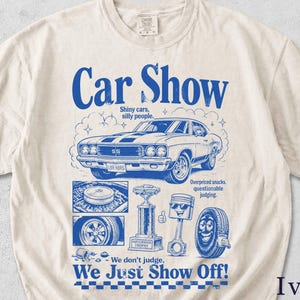 May include: Ivory-colored t-shirt with a blue graphic that reads "Car Show." The design includes a classic car, car parts, and the text "We don't judge, We Just Show Off!"