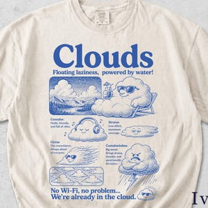 May include: Ivory-colored t-shirt with a blue graphic design featuring the word "Clouds" and various cloud illustrations. The text "Floating laziness, powered by water!" is also included. The shirt has a crew neck and short sleeves.