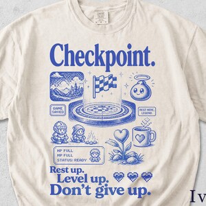 Può includere: T-shirt color avorio con un design grafico blu con la parola "Checkpoint" e varie illustrazioni a tema gaming. Il design include una bandiera a scacchi, personaggi pixelati e le frasi "Rest up", "Level up" e "Don't give up."
