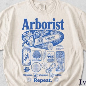 Könnte beinhalten: Elfenbeinfarbenes T-Shirt mit blauem Grafikdesign. Das Design zeigt das Wort "Arborist" über einer Kettensägenillustration und dem Text "Sawdust Therapy". Zusätzliche Illustrationen umfassen Bäume, Blätter und Kletterausrüstung. Der Text "Climbing. Chopping. Coffee. Repeat." ist ebenfalls enthalten.
