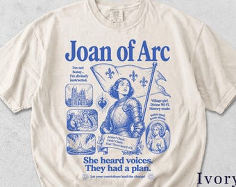 Könnte beinhalten: Elfenbeinfarbenes T-Shirt mit einem blauen Aufdruck von Jeanne d'Arc, die eine Fahne hält. Das Design enthält den Text "Joan of Arc", "She heard voices. They had a plan" und andere Phrasen. Das T-Shirt hat einen Rundhalsausschnitt und kurze Ärmel.