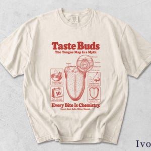 May include: An ivory short-sleeve t-shirt featuring a red graphic print of a tongue anatomy diagram. The text reads Taste Buds, The Tongue Map Is a Myth, and Every Bite Is Chemistry, along with labels for Sweet, Sour, Salty, Bitter, and Umami.