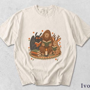 Puede incluir: Una camiseta de manga corta color marfil con un estampado gráfico de una polilla, un sasquatch y un jackalope sentados sobre una alfombra estampada. Las criaturas leen libros a la luz de las velas, rodeadas de pequeñas flores naranjas y estrellas.
