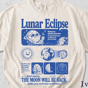 Könnte beinhalten: Ein elfenbeinfarbenes Kurzarm-T-Shirt mit einem blauen Grafikdruck mit dem Titel Lunar Eclipse. Das Design enthält Cartoon-Illustrationen von Mond, Sonne und Erde mit Texten wie Nature's shade thrower, Earth's way of putting on a show und Don't worry, THE MOON WILL BE BACK. Unlike your motivation on Mondays.