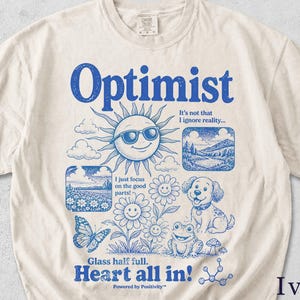 May include: An ivory t-shirt featuring a blue graphic print. The text reads Optimist, It's not that I ignore reality, I just focus on the good parts, Glass half full, Heart all in, and Powered by Positivity. The design includes a sun with sunglasses, flowers, a dog, a frog, a butterfly, and landscape scenes.