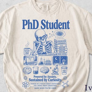 Op de afbeelding: Een ivoorkleurig t-shirt met een blauwe afbeelding van een skelet met een bril aan een bureau. De tekst luidt PhD Student, Caffeine Fuels Hope, Powered by Anxiety, Sustained by Curiosity en Sleep When You're Hired, naast diverse academische iconen.