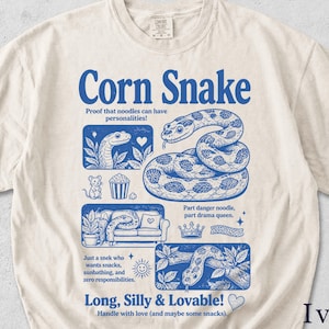 Op de afbeelding: Ivoor t-shirt met een blauw grafisch ontwerp met de tekst "Corn Snake". Het ontwerp bevat illustraties van slangen, een muis en de tekst "Long, Silly & Lovable!" en "Handle with love (and maybe some snacks)."