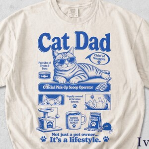 May include: Ivory-colored t-shirt with a blue graphic that reads "Cat Dad." The graphic features a cat wearing sunglasses, along with other cat-related images and text. The shirt is made of a soft material.