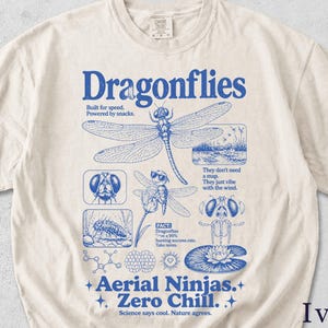 Könnte beinhalten: Ein elfenbeinfarbenes T-Shirt mit einem blauen Grafikdruck von Libellen. Das Design enthält den Text "Dragonflies," "Built for speed. Powered by snacks," "Aerial Ninjas. Zero Chill," und "Science says cool. Nature agrees," sowie verschiedene Libellenillustrationen und wissenschaftliche Fakten.