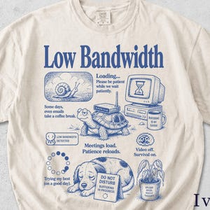 May include: An ivory short-sleeve t-shirt featuring a blue graphic print with the text Low Bandwidth. The design includes illustrations of a snail, a turtle with a router, a computer monitor with an hourglass, a coffee mug, and a sleeping dog.