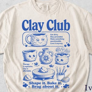 Könnte beinhalten: Elfenbeinfarbenes T-Shirt mit blauem "Clay Club"-Aufdruck. Das Design zeigt Illustrationen von Töpferwaren wie einer Tasse, einer Vase und einem Pflanzgefäß sowie den Text "Shape it. Bake it. Brag about it."