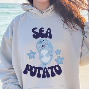 Może przedstawiać: Jasnobrązowa bluza z kapturem z przednią kieszenią i kapturem ze sznurkiem. Z przodu widnieje napis "SEA POTATO" w ciemnoniebieskim kolorze, z ilustracją kreskówki manata i niebieskimi kwiatowymi akcentami. Bluza z kapturem jest noszona na plaży.