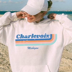 May include: White hoodie with the word "Charlevoix" in blue, orange, and teal retro font, with "Michigan" below. The hoodie has a front pocket and is worn with a white cap and black shorts. The background is a beach with blue water.