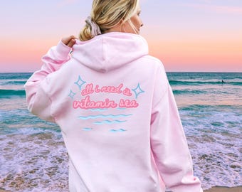 May include: Pink hoodie with the text "all i need is vitamin sea" in pink and blue lettering with a wave design and stars on the back. The hoodie is being worn by a person standing on a beach.