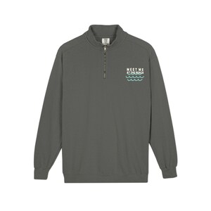 May include: A charcoal gray long-sleeve quarter-zip sweatshirt featuring a small white graphic on the left chest that reads "MEET ME AT THE BEACH" above three light teal wavy lines representing ocean waves, with a high collar and zipper.