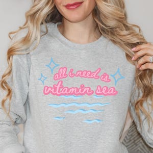 Könnte beinhalten: Ein grauer Pullover mit dem Text "all i need is vitamin sea" in rosa Schrift mit blauen Sternen und Wellenlinien.