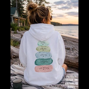 May include: A white hooded sweatshirt featuring a vertical stack of five watercolor-style brushstrokes on the back. Each stroke contains the text: Leland, Glen Lake, Torch Lake, Charlevoix, and Lake Michigan, each accompanied by a small heart icon.