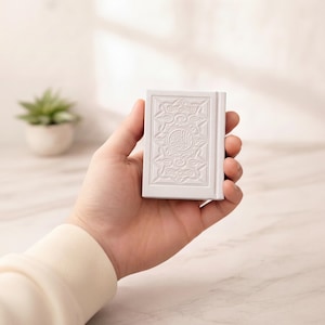 May include: A small, white book with embossed patterns on the cover, held in a hand. The cover has a central circular emblem with Arabic script and floral designs. The background is a soft, neutral colour.