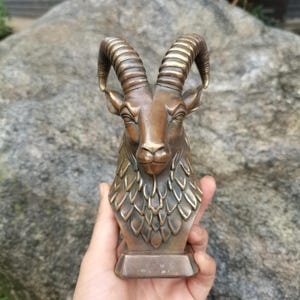 Puede incluir: Una escultura de tono bronce de una cabeza de carnero con cuernos grandes, curvos y estriados, y un área de cuello detallada con texturas superpuestas en forma de hoja. El busto está montado sobre una base rectangular con acabado metálico.
