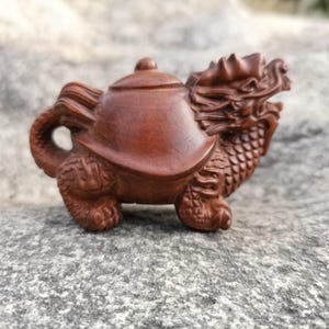 Puede incluir: Una detallada escultura de madera de color marrón rojizo de una criatura mítica. La escultura combina la forma de una tetera con un dragón, con intrincadas tallas y una superficie texturizada. La cabeza del dragón es visible, y la tetera tiene tapa y asa.