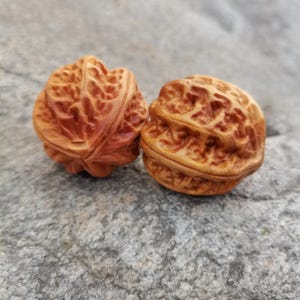 Puede incluir: Dos nueces de madera talladas intrincadamente, que muestran patrones y texturas detalladas. Las nueces son de color marrón cálido, sobre una superficie de piedra gris. Los grabados resaltan la forma natural de las nueces.