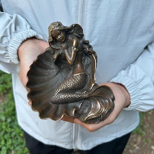 Puede incluir: Una escultura de tono bronce de una sirena con cabello largo y fluido, sentada dentro de una concha grande y abierta. La figura presenta escamas detalladas en la cola y una base de concha texturizada, con un acabado metálico y reflejos oscuros.
