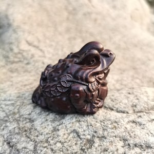 Puede incluir: Una detallada talla de madera marrón oscuro de un sapo, con intrincados detalles y una moneda en la boca. El sapo está posicionado sobre una superficie de piedra gris clara texturizada, mostrando la artesanía de la figura.