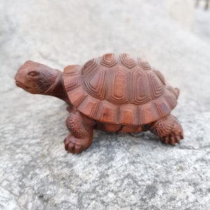 May include: A detailed wooden turtle figurine, carved from a reddish-brown wood. The turtle has a textured shell with geometric patterns and detailed legs and head. The figurine is displayed on a textured, light gray surface.