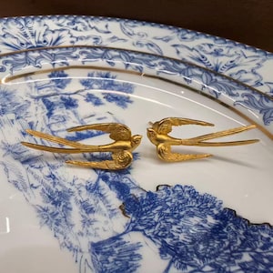 May include: A pair of gold-toned metal swallow bird earrings featuring detailed wing and tail feather textures. Each bird is depicted in a flying position with wings extended, showcasing a metallic finish and intricate engraved lines on the surface.