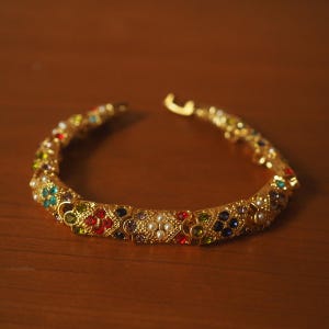 Op de afbeelding: Een goudkleurige ketting met kleurrijke edelstenen en kleine witte parels. De ketting heeft decoratieve segmenten, elk versierd met een verscheidenheid aan gekleurde stenen. De sluiting is zichtbaar aan het einde van de ketting.