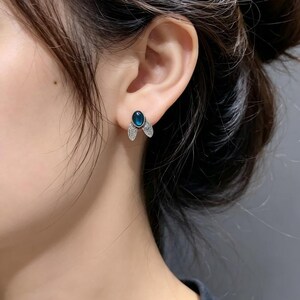 May include: A stud earring featuring a central oval-shaped dark blue stone set in a silver-toned metal frame. The design includes two small, textured silver-toned leaf-like accents extending downward from the base of the central blue stone.
