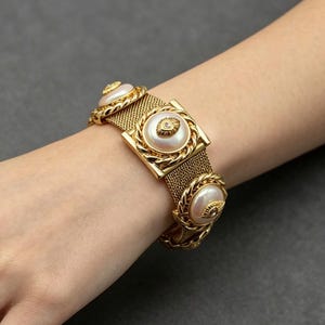 Puede incluir: Pulsera dorada con una banda de malla y elementos decorativos. La pulsera tiene tres grandes acentos redondos con forma de perla y una pieza central cuadrada. La pulsera mide aproximadamente 17,78 cm de largo.