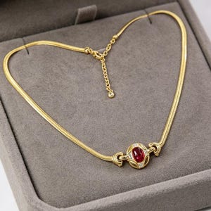 May include: A gold necklace featuring a central oval ruby gemstone, framed by small white stones. The necklace is displayed in a grey jewellery box, highlighting its elegant design and craftsmanship.