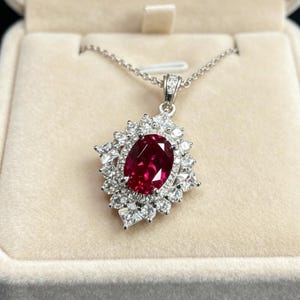 May include: A silver necklace with a large, oval, ruby-red gemstone pendant. The pendant is surrounded by a halo of smaller, clear gemstones. The necklace is displayed in a beige jewelry box.