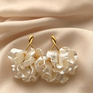 Op de afbeelding: Een paar elegante oorbellen met een cluster van witte, bloembladachtige vormen. De oorbellen hebben goudkleurige pinnen en accenten, wat een verfijnd en vrouwelijk ontwerp creëert. Ze staan tegen een zachte, crèmekleurige achtergrond.