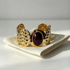 Puede incluir: Un brazalete tipo puño de color dorado con acabado pulido y un patrón de panal calado. El centro del brazalete está adornado con una piedra cabujón grande, lisa y de forma ovalada en color burdeos oscuro, engastada en un marco dorado.