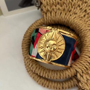Könnte beinhalten: Goldfarbenes Metallarmband mit einem Sonnenmotiv. Das Armband hat ein farbenfrohes Stoffband mit roten, blauen, grünen und weißen Mustern. Das Armband liegt auf einem geflochtenen Seil.