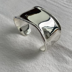 Op de afbeelding: Een brede, gepolijste zilveren armband met een glad, reflecterend oppervlak en een licht onregelmatige, organische vorm. Het metaal heeft een hoogglanzende afwerking die het licht opvangt en zachte rondingen en contouren creëert.