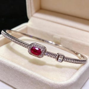 Peut inclure: Un bracelet jonc argenté avec une pierre précieuse rouge rubis rectangulaire au centre, entourée d'un halo de petits diamants transparents. Le bracelet comporte également une petite pierre précieuse transparente rectangulaire. Présenté dans un écrin blanc.