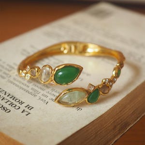 以下が含まれることがあります： 緑と透明な宝石のアクセントが付いた、金色のブレスレット。 オープンカフのデザインは、涙の形をした緑色の石と、より小さく丸い透明な石が特徴です。 ブレスレットはイタリア語のテキストが書かれた本の上に置かれています。