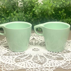 May include: Two light green mugs with a handle, set on a white lace doily. The mugs have a simple, classic design and a smooth, glossy finish. The background features green foliage.