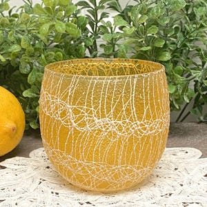 May include: A yellow glass votive candle holder with a white web-like design. The candle holder is round and sits on a white lace doily. A lemon is visible on the left side of the image, with green foliage in the background.