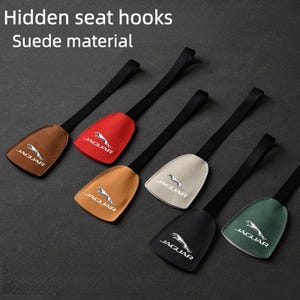 May include: Hidden seat hooks made of suede material. The image shows six seat hooks in various colors: brown, red, tan, off-white, black, and green. Each hook has a black strap and a Jaguar logo.