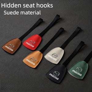 Könnte beinhalten: Sechs Autositzhaken in verschiedenen Farben: Braun, Rot, Beige, Off-White, Schwarz und Grün. Jeder Haken hat einen schwarzen Riemen und eine Wildleder-Oberfläche mit dem Mazda-Logo. Der Text oben lautet "Hidden seat hooks" und "Suede material."