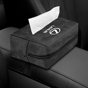 Puede incluir: Caja de pañuelos negra con el logotipo de Lexus en blanco. La caja rectangular tiene cierre de cremallera y una pequeña lengüeta. Los pañuelos blancos sobresalen de la parte superior. La caja está colocada en la consola central de un coche.