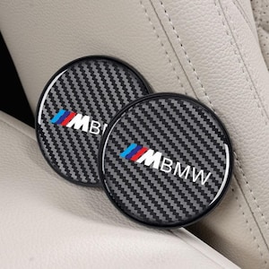 May include: Two round car coasters with a carbon fiber pattern and a black border. The coasters feature the BMW logo in white, blue, and red. They are resting on a light gray car seat.