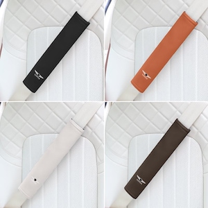 May include: Four seat belt shoulder pads in black, tan, cream, and brown. Each pad has a Genesis logo. The pads are shown on a white car seat with a quilted pattern. These accessories are designed to provide comfort and protection.