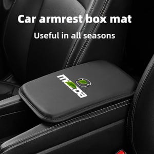 Op de afbeelding: Zwarte auto-armsteun box mat met witte stiksels. De mat heeft het Mazda-logo en de tekst "Useful in all seasons". De mat is ontworpen om in een auto te passen.