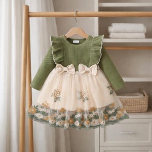 May include: A long-sleeved olive green dress with a cream-colored tulle skirt. The dress features three cream-colored bows at the waist and floral embroidery along the skirt's hem. The dress is hanging on a wooden hanger.