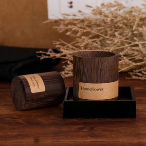 May include: Two dark brown wooden reed diffusers, one cylindrical and the other a cylinder with a label that reads "HomeFlower". The cylindrical diffuser is on a black square tray. The other diffuser has a label that reads "Reed Diffuser Since 2009".